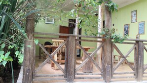 A rustic wooden fence with a stone base, a wooden table and chairs in the background, and a green wall with framed pictures. - Kibale Eco Home  (Kibale)