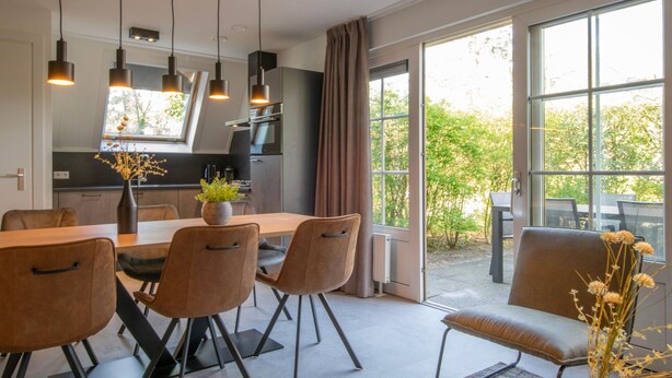 A modern dining area with a large table and chairs, a vase with flowers, and a view of the outdoors through large windows. - Holiday Home Near Biesbosch & Breda (Oosterhout)