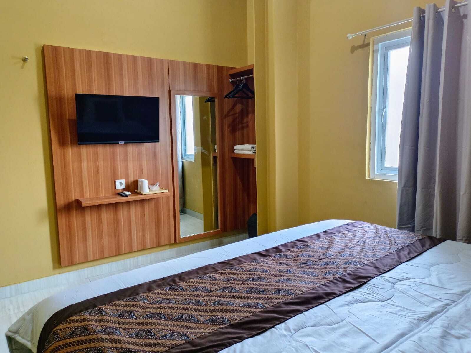 A hotel room with a bed, a TV mounted on a wooden panel, a mirror, and a window with curtains.