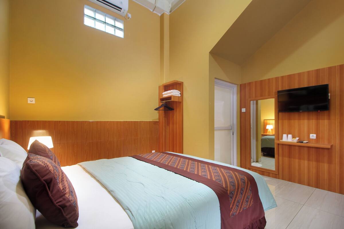 A hotel room with a bed, a wooden headboard, a TV mounted on the wall, a mirror, and a small shelf with toiletries.