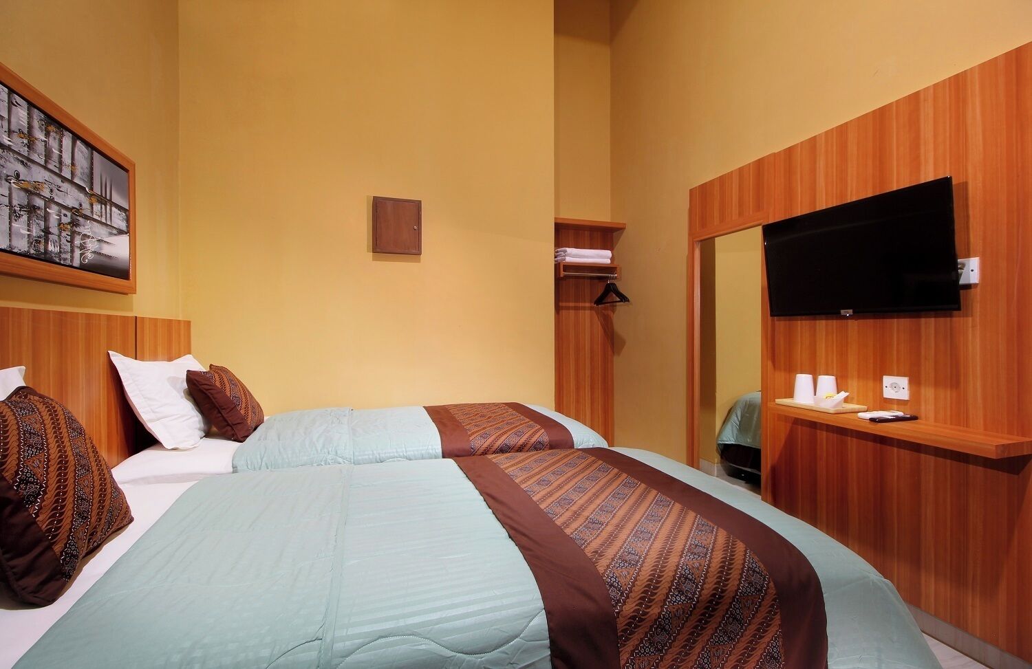 A hotel room with two beds, a wooden wardrobe, a television, and a small table with cups and a remote control.