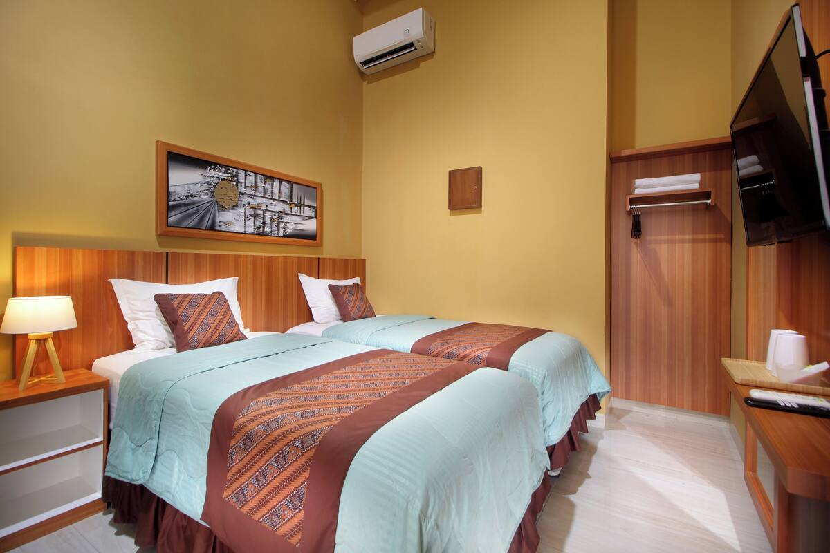 A hotel room with two beds, a wooden headboard, a mounted TV, a wall-mounted air conditioner, and a framed picture above the beds.