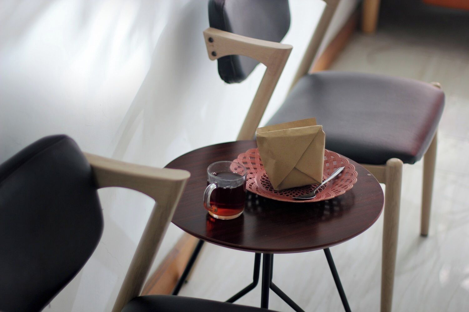 A small round table with a dark wooden top, a clear glass mug with a beverage, a folded brown paper bag, and a silver spoon on a red doily.