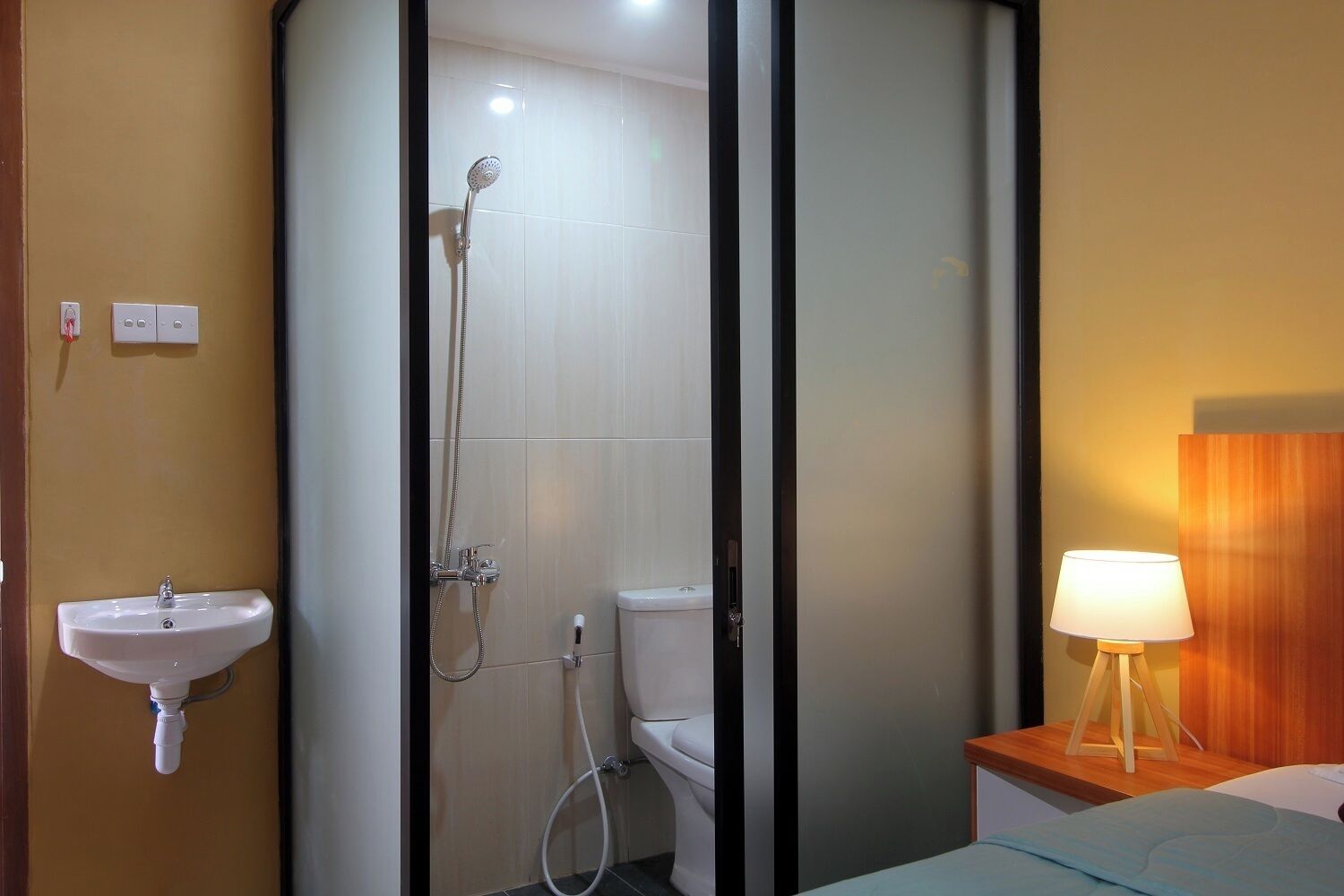 A hotel bathroom with a shower, toilet, sink, and bedside table with a lamp.