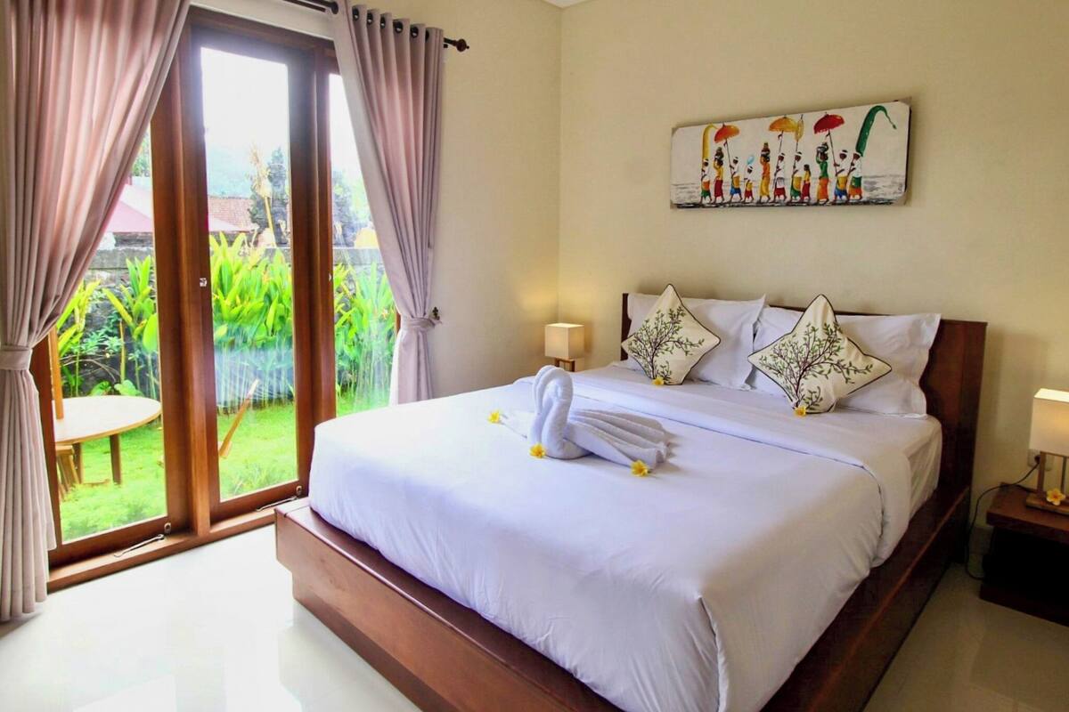 A bedroom with a bed, curtains, a painting, and a view of greenery.