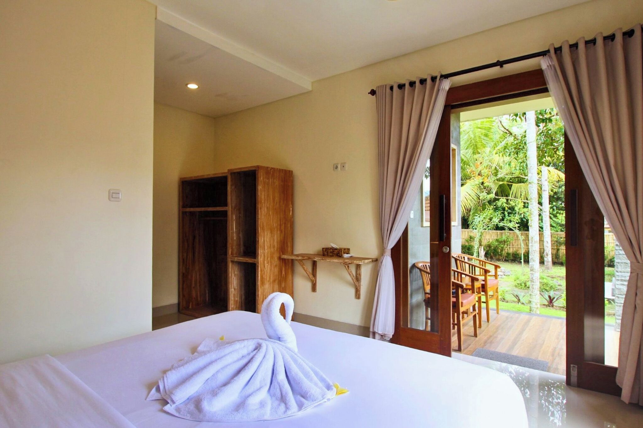 A bedroom with a bed, a wooden wardrobe, a small table, and a view of an outdoor area with a patio and chairs.