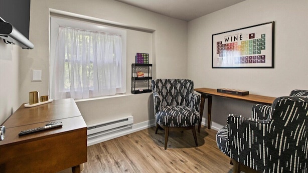 Una habitación con piso de madera, una ventana con cortinas traslúcidas, una carta de vinos enmarcada en la pared, un sillón con estampado en blanco y negro, y una pequeña mesa con un control remoto sobre un escritorio de madera.