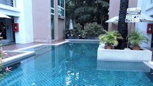 Una piscina con suelo de baldosas, rodeada de plantas en macetas y un edificio con un cartel.