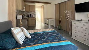Un dormitorio con cama, televisor, cómoda y una kitchenette al fondo.