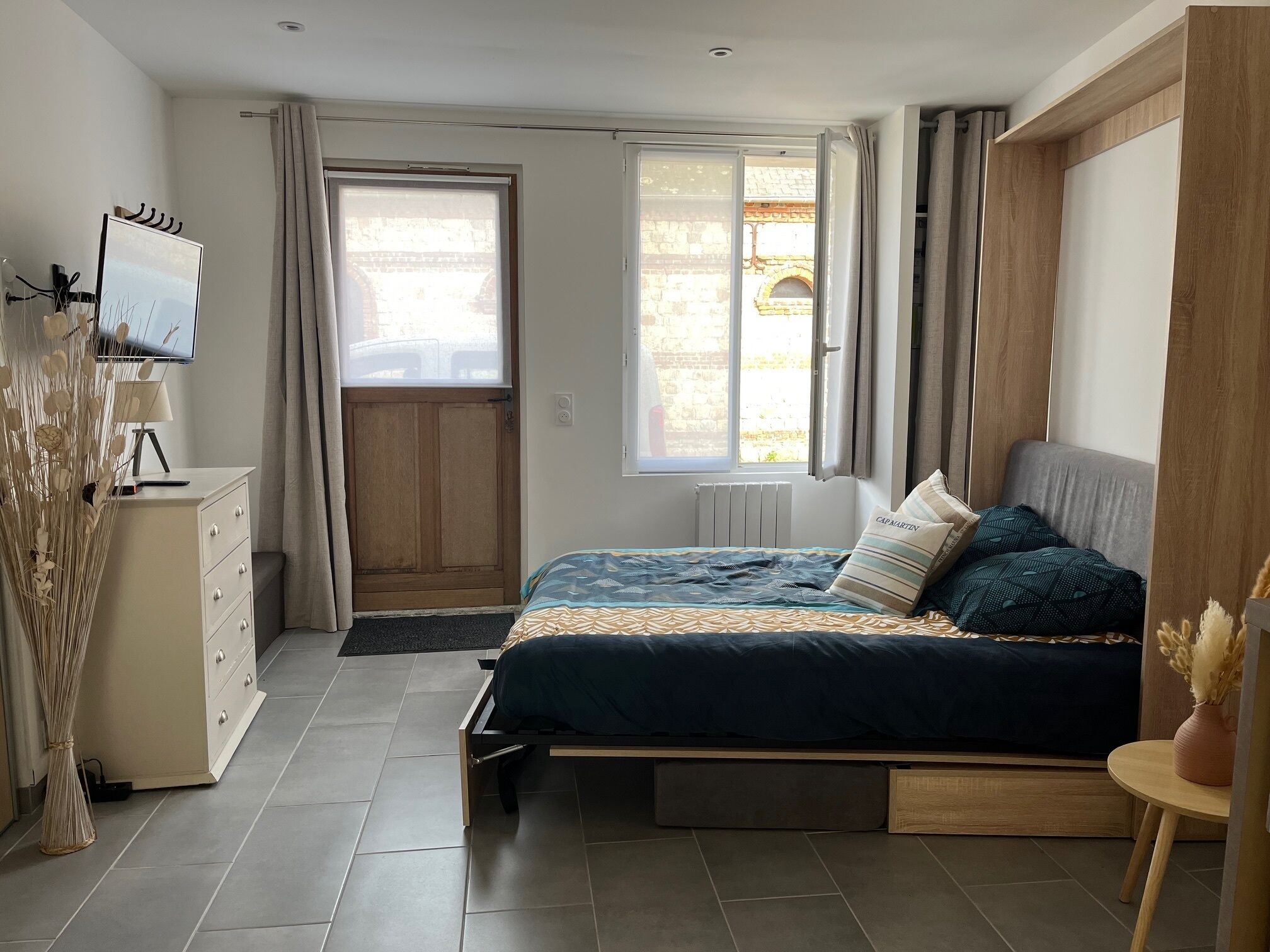 Un dormitorio con cama, televisor, armario de madera y ventana con cortinas.