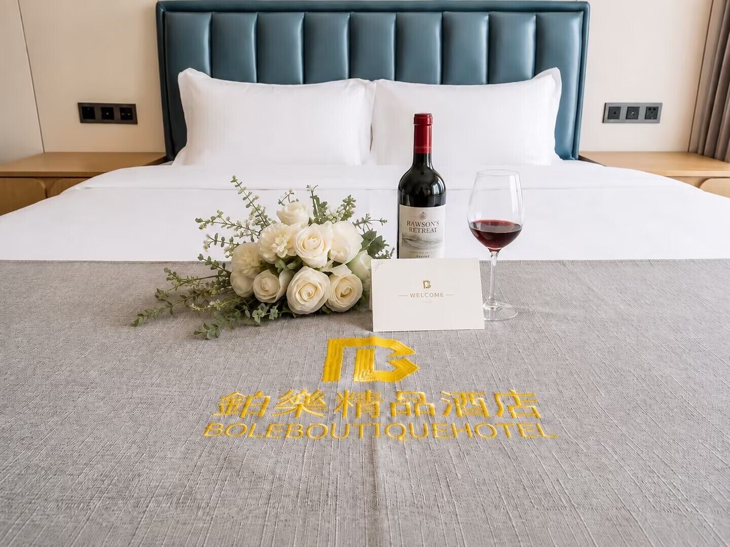 Una camera d'albergo con un letto, un bouquet di rose bianche, una bottiglia di vino e un biglietto di benvenuto.