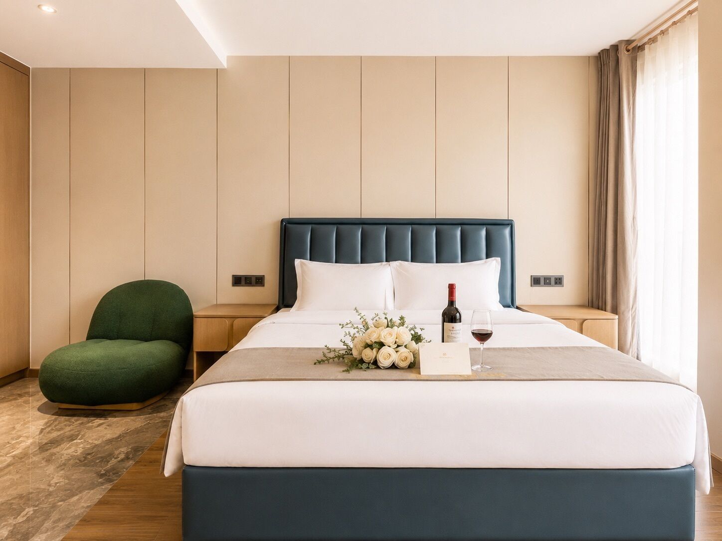 Une chambre d’hôtel avec un lit, un fauteuil vert, une table de chevet et une petite table de nuit sur laquelle se trouvent une bouteille de vin et un verre.