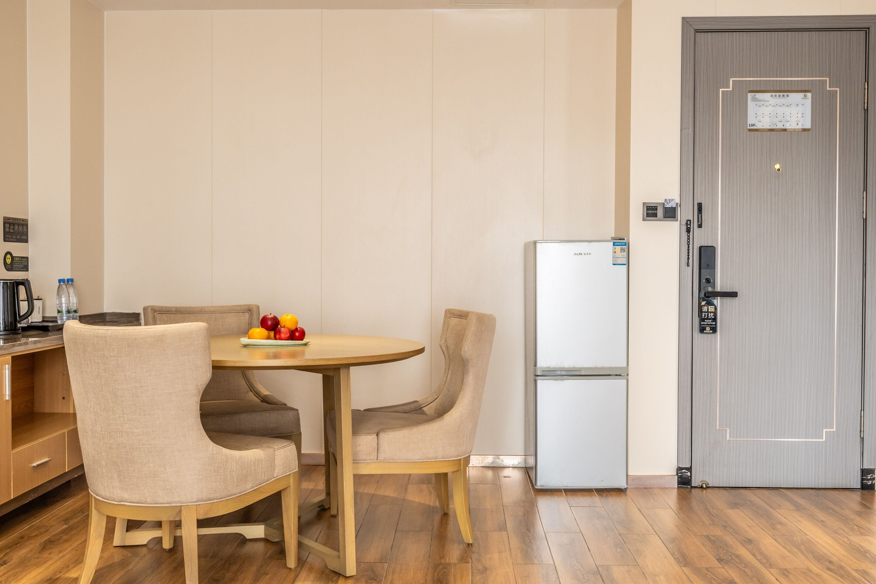 A compact hotel room with a wooden dining table, two chairs, a refrigerator, and a door with a lock and a small sign.