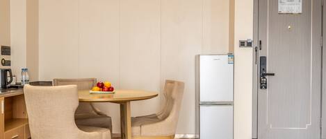 A compact hotel room with a wooden dining table, two chairs, a refrigerator, and a door with a lock and a small sign.