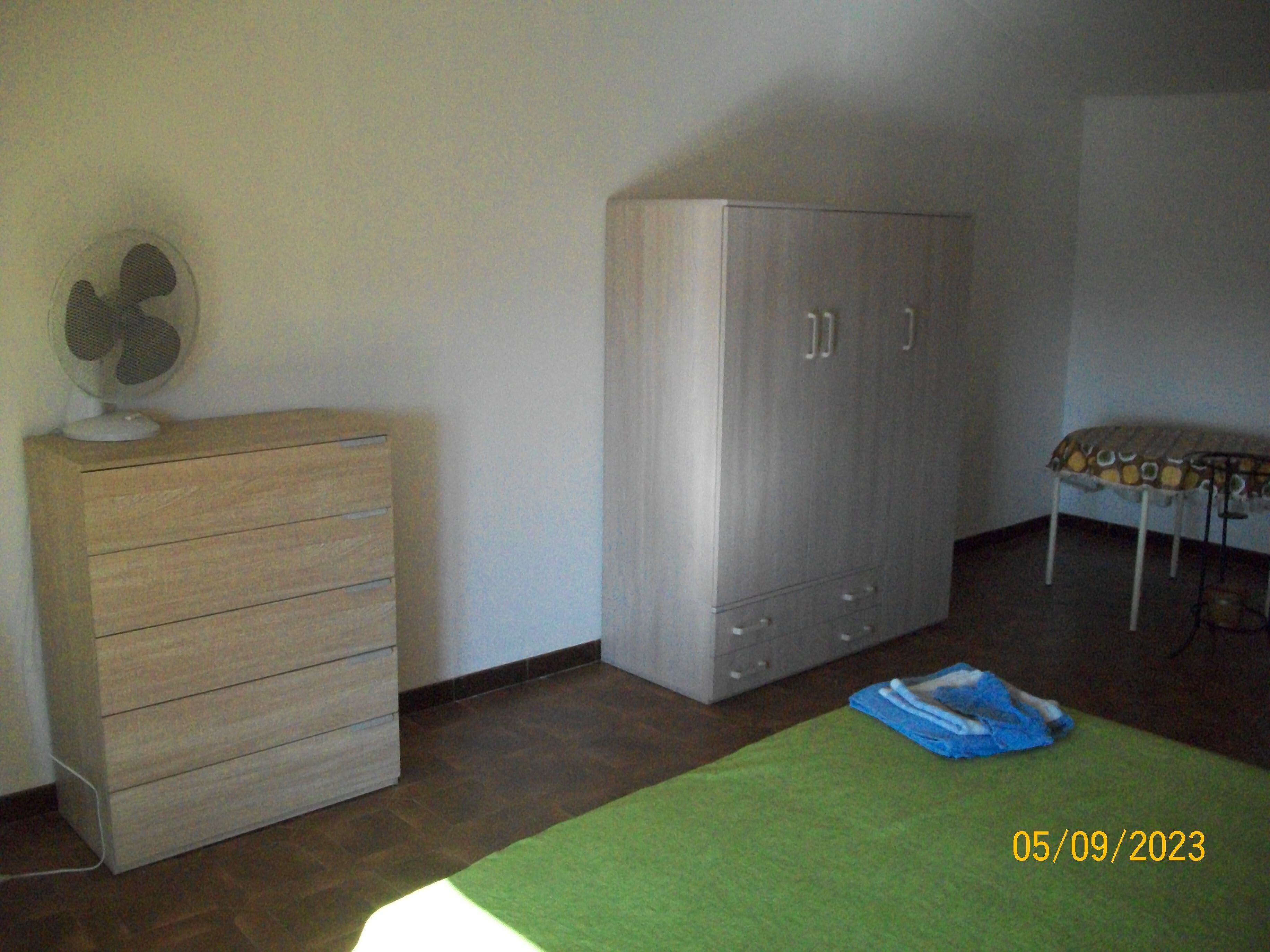 Una camera da letto con un comò in legno, un armadio grigio, una coperta verde e una ventola sul comò.