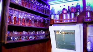A bar area with shelves of glasses and bottles, a refrigerator, and a view of the water outside. - AMAZING in Ortigia  (Siracusa)