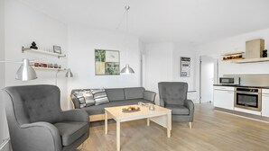 Una sala de estar moderna con un sofá gris, dos sillones, una mesa de centro y una zona de cocina con microondas y horno.