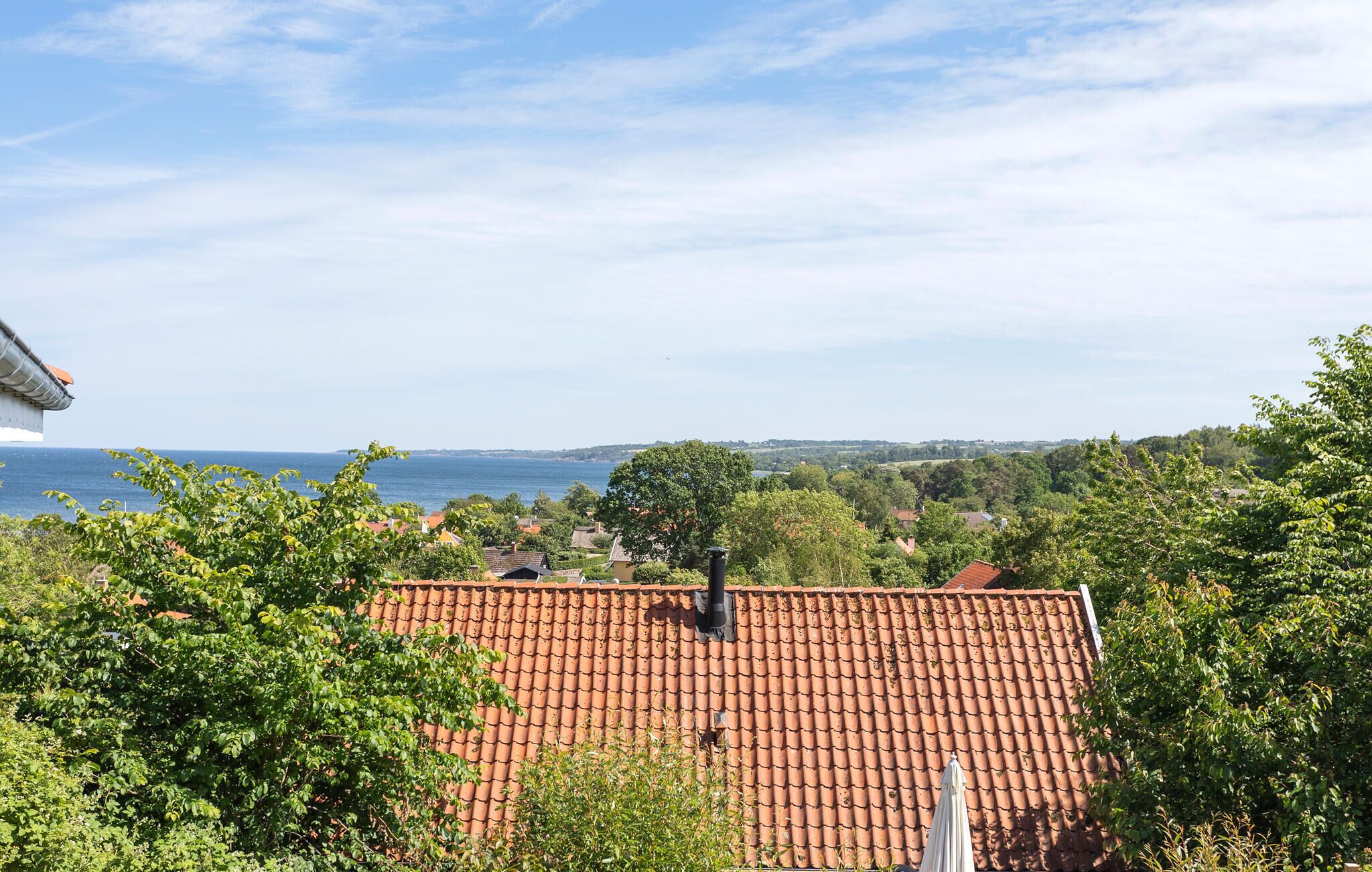 A red-tiled roof house with a chimney, surrounded by greenery and overlooking a body of water.