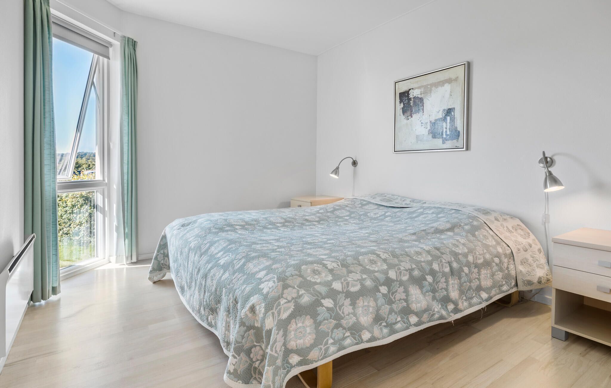 Een slaapkamer met een bed, nachtlampjes, een nachtkastje, een raam met gordijnen en een aan de muur hangend schilderij.