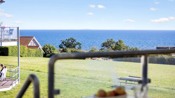 A view of a grassy area with a bench and a table, overlooking a body of water and a building with a red roof. - Gudhjem Søpark Lejl. 12 (Gudhjem)