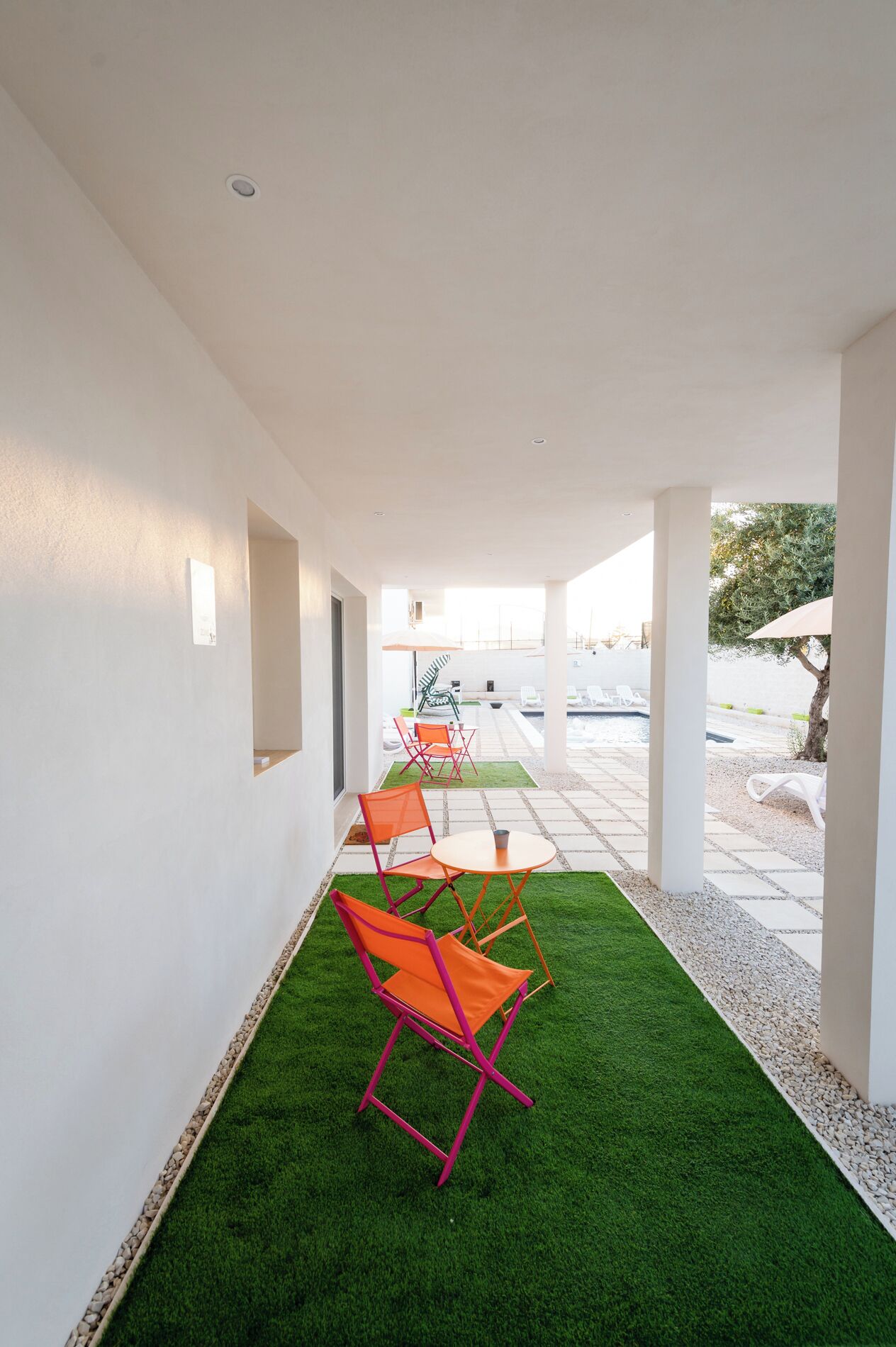 Ein Korridor mit künstlichem Gras, der zu einem Poolbereich führt, mit einer Reihe orangefarbener Stühle und einem weißen Sonnenschirm.