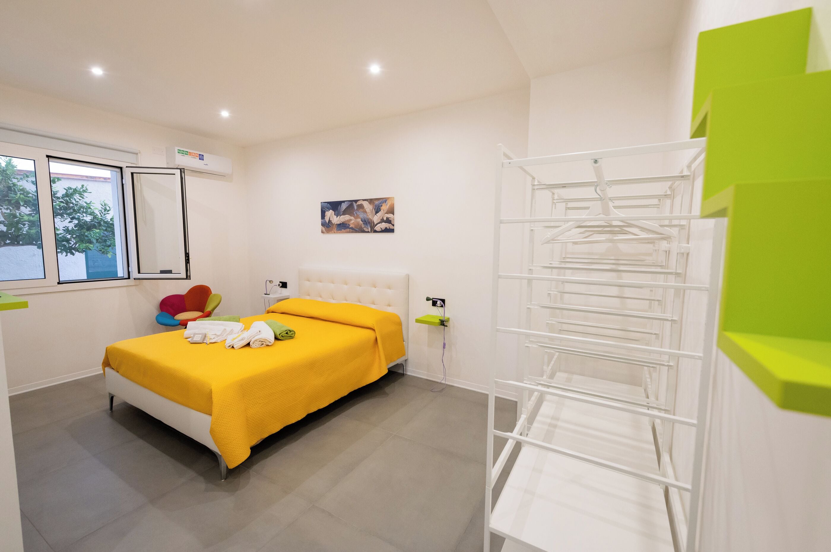 Un dormitorio con cama, ventana, aire acondicionado de pared, un cuadro y un estante blanco.