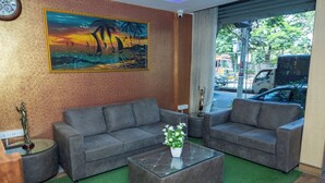 A living room with two grey sofas, a glass coffee table with a white vase, a painting of a sunset, and a view of the street outside. - Ishwerya Suites (Bengaluru)