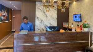 A hotel reception area with a marble wall, a receptionist, a desk with a computer, a television, and a non-smoking sign. - Ishwerya Suites (Bengaluru)