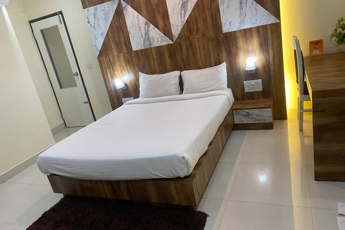 A modern bedroom with a wooden bed frame, white bedding, a dark rug, and a wall-mounted light fixture.
