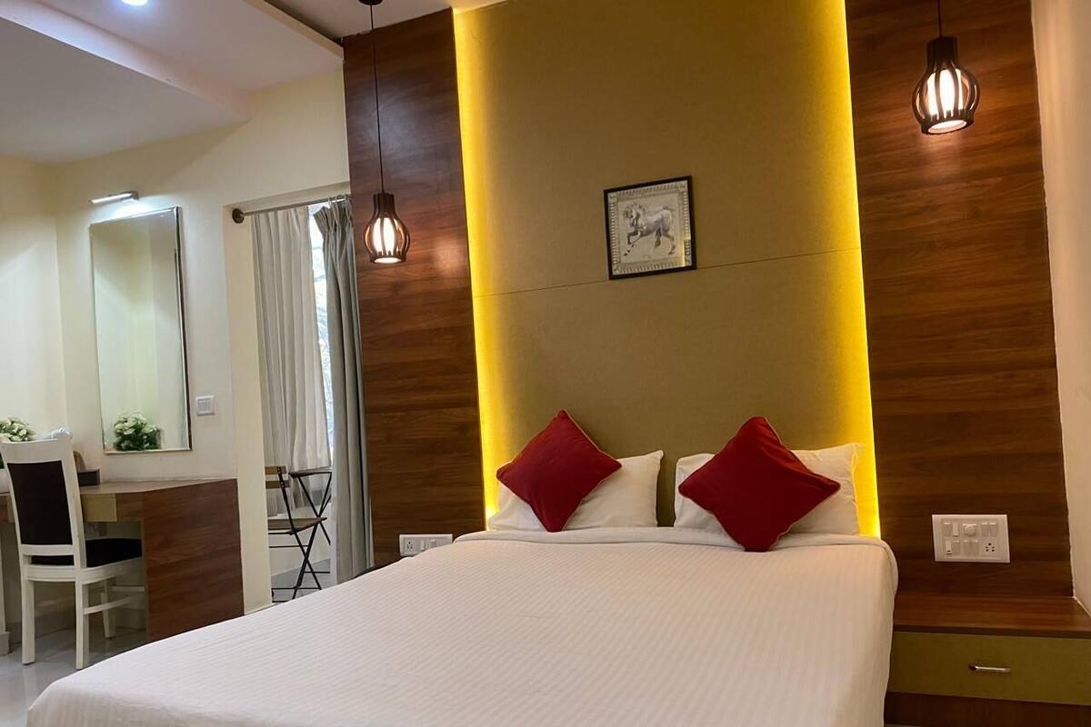 A hotel room with a large bed, a desk, and a chair. There is a window with curtains, a wall-mounted light fixture, and a power outlet.