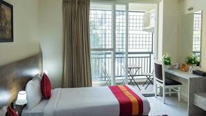 A hotel room with a bed, a desk, a chair, a window with curtains, and a balcony with a railing. - Ishwerya Suites (Bengaluru)