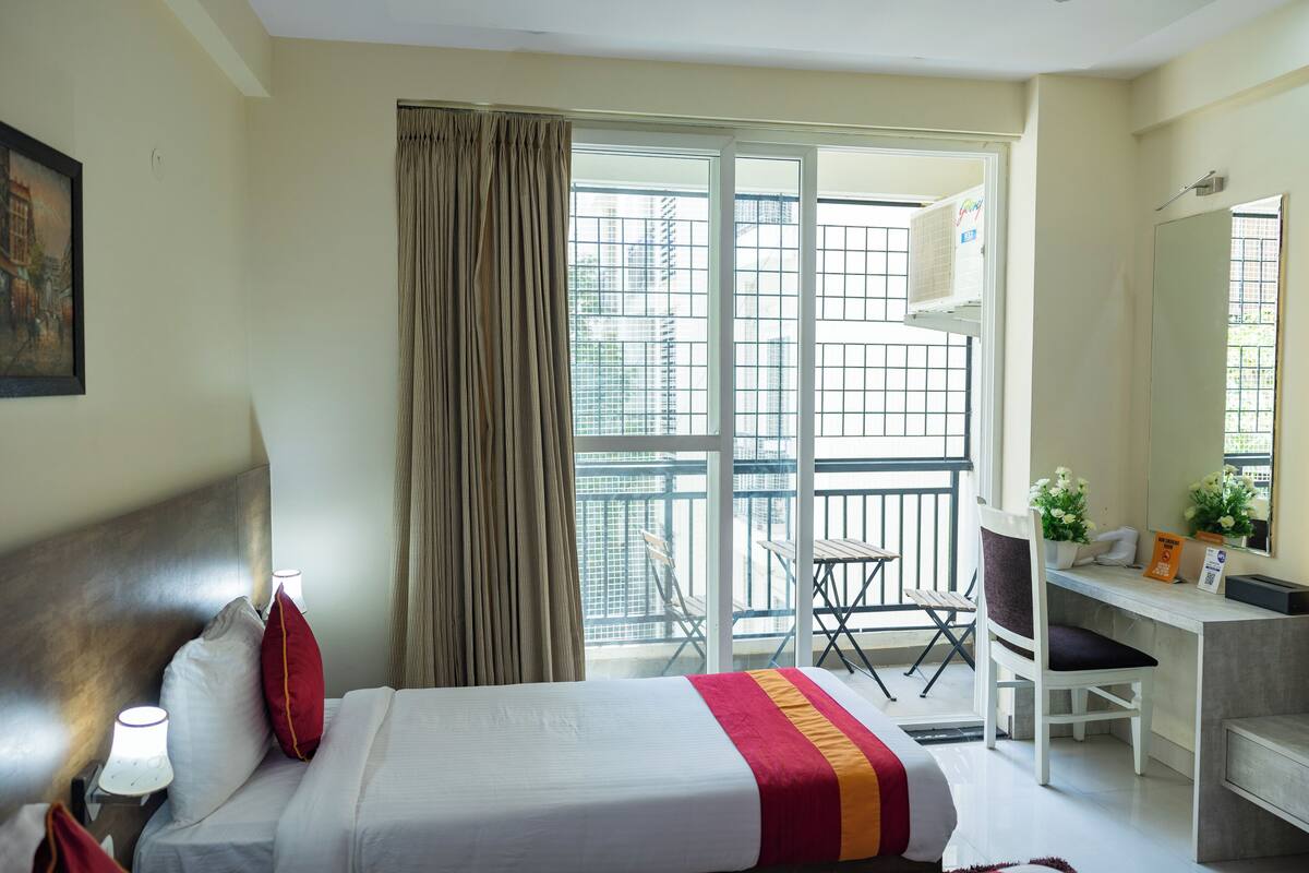 A hotel room with a bed, a desk, a chair, a window with curtains, and a balcony with a railing.