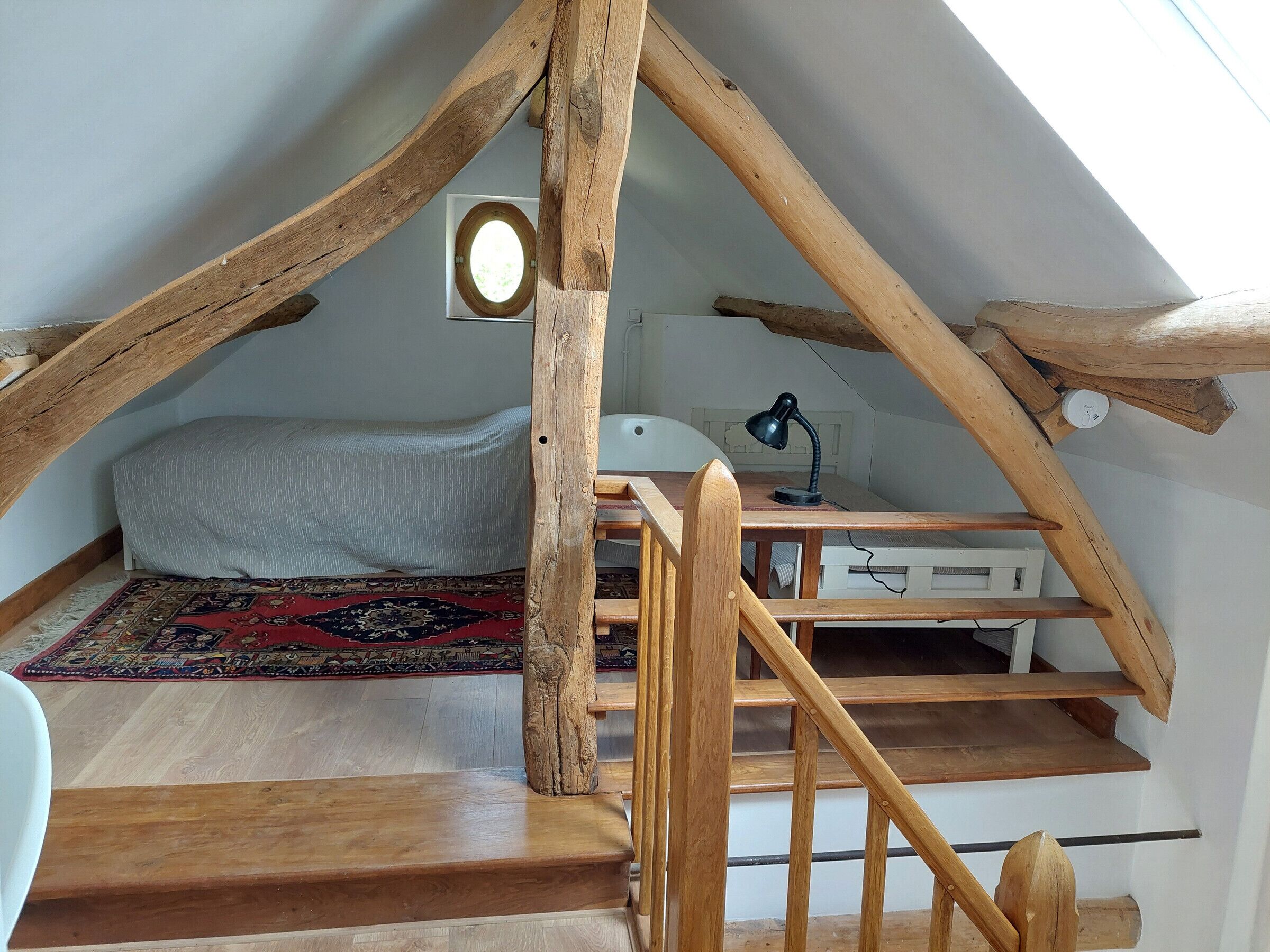A loft bedroom with a sloped ceiling, a bed, a desk, a chair, a small table, a lamp, a radiator, and a staircase.