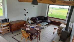 Un salón acogedor con una mesa de madera para cuatro personas, un sofá de color oscuro y un amplio ventanal con vistas a un campo verde.