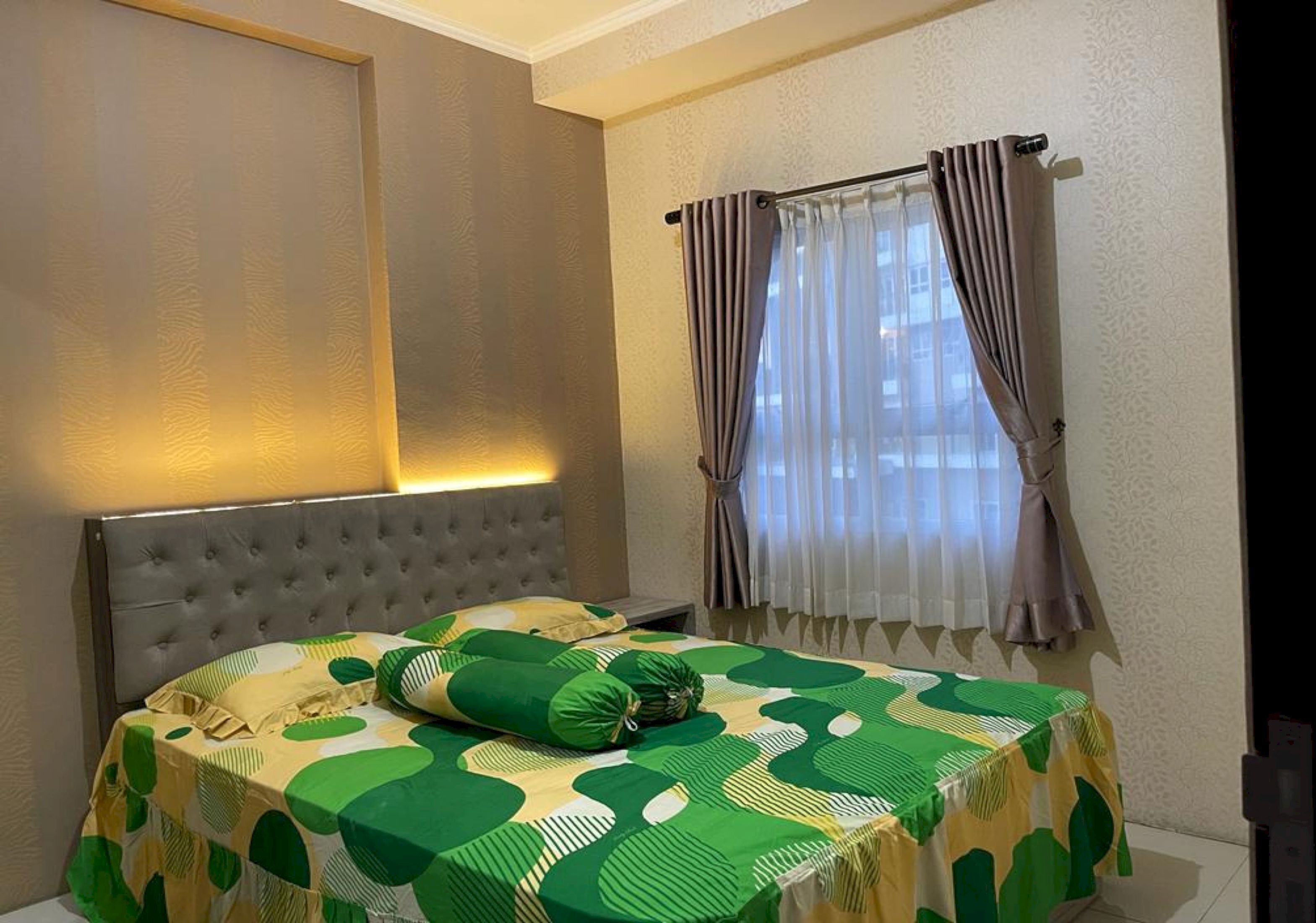 A bedroom with a tufted headboard, a bed with a green and yellow patterned bedspread, and a window with curtains.