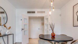 A dining area with a round table, four chairs, a vase with dried flowers, and a hanging light fixture. - Luxury 2BR Apt in Heart of Puerto Banús – Steps from Beach with Mountain Views (Marbella)