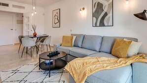 A modern living room with a grey sofa, a round coffee table, and a dining area with chairs and a table. - Luxury 2BR Apt in Heart of Puerto Banús – Steps from Beach with Mountain Views (Marbella)