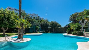 A large, clear blue swimming pool surrounded by palm trees and greenery, with a building in the background. - Luxury 2BR Apt in Heart of Puerto Banús – Steps from Beach with Mountain Views (Marbella)