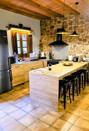 A modern kitchen with a central island, black bar stools, a stone wall, and a wooden ceiling. - More Morrenyà - Garrotxa Country House (Sant Jaume de Llierca)