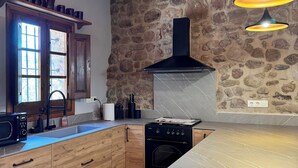 A kitchen with a stone wall, wooden cabinets, a black stove, and a window with a view. - More Morrenyà - Rural House Garrotxa (Sant Jaume de Llierca)