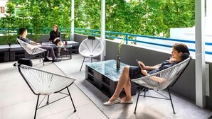 Un espace extérieur aménagé avec des chaises modernes et une table à plateau de verre, entouré de verdure et d’une rambarde bleue.