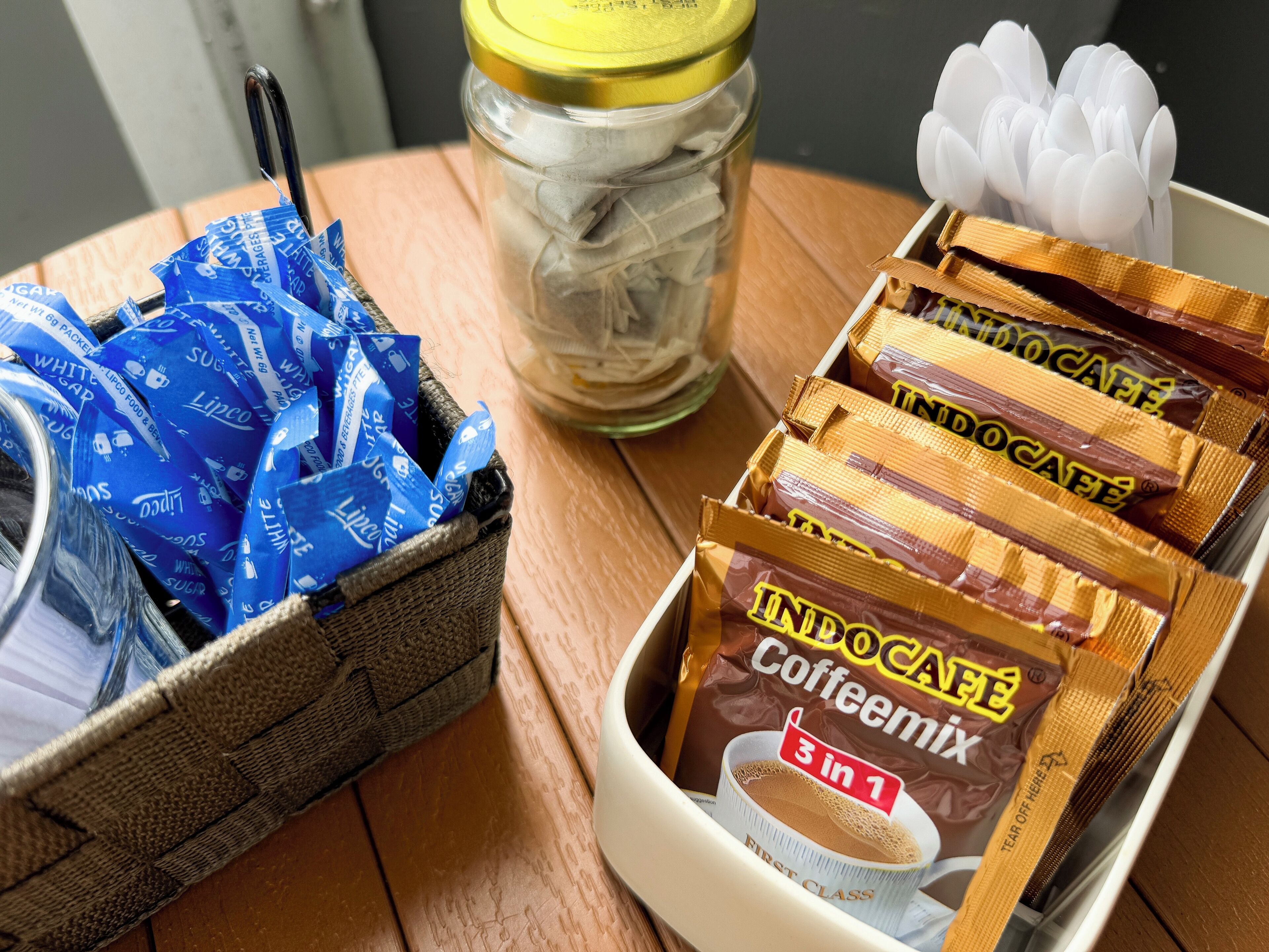 A wooden table with a basket containing blue Lipco packaging, a jar with a yellow lid, and a tray of Indocafé Caffeine packets.