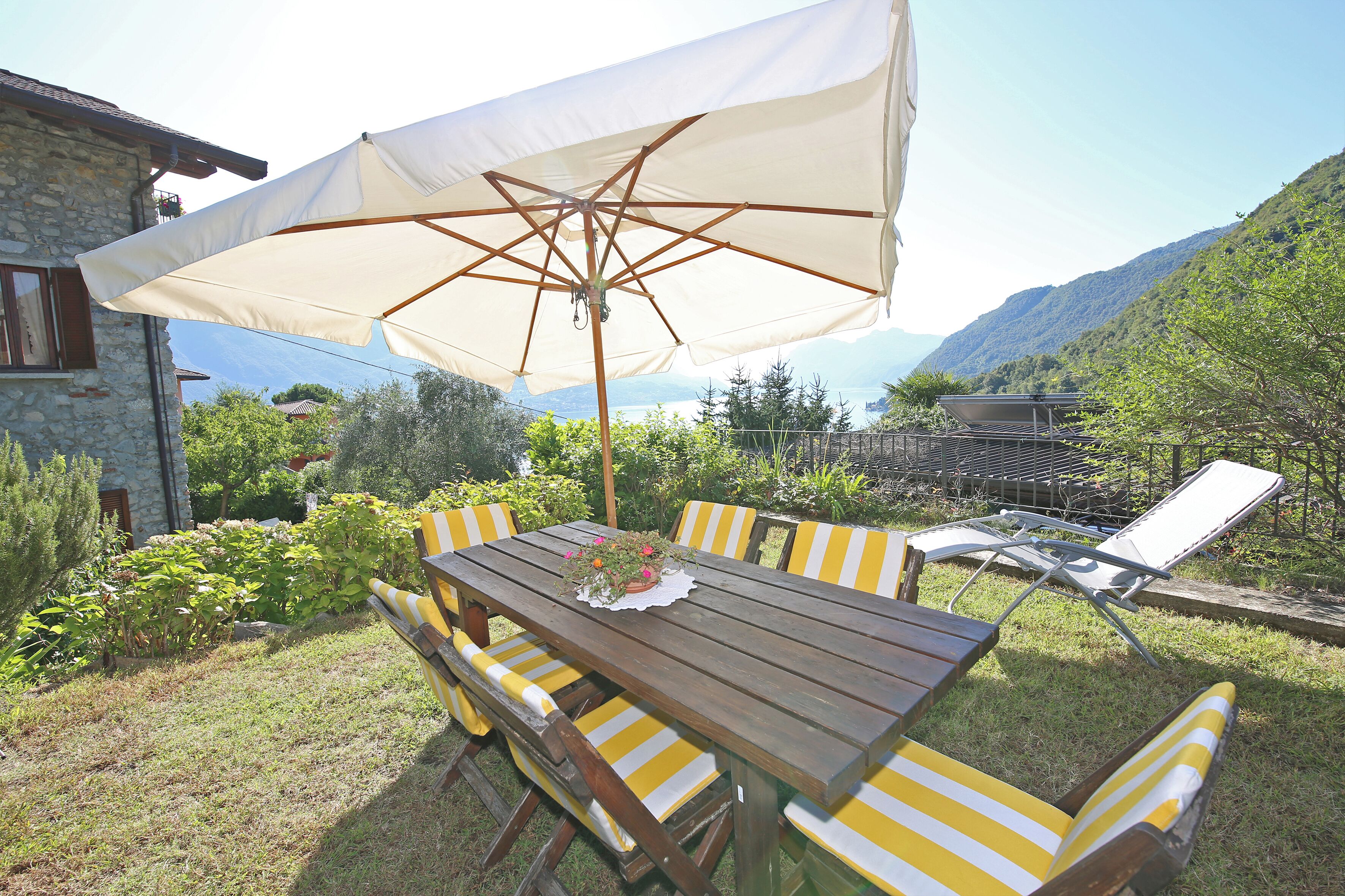 Een terras met een houten tafel en stoelen met gele strepen, een parasol en uitzicht op bergen en een meer.