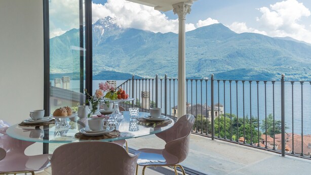 Zona pranzo con tavolo in vetro e sedie rosa, con vista su un lago e montagne.
