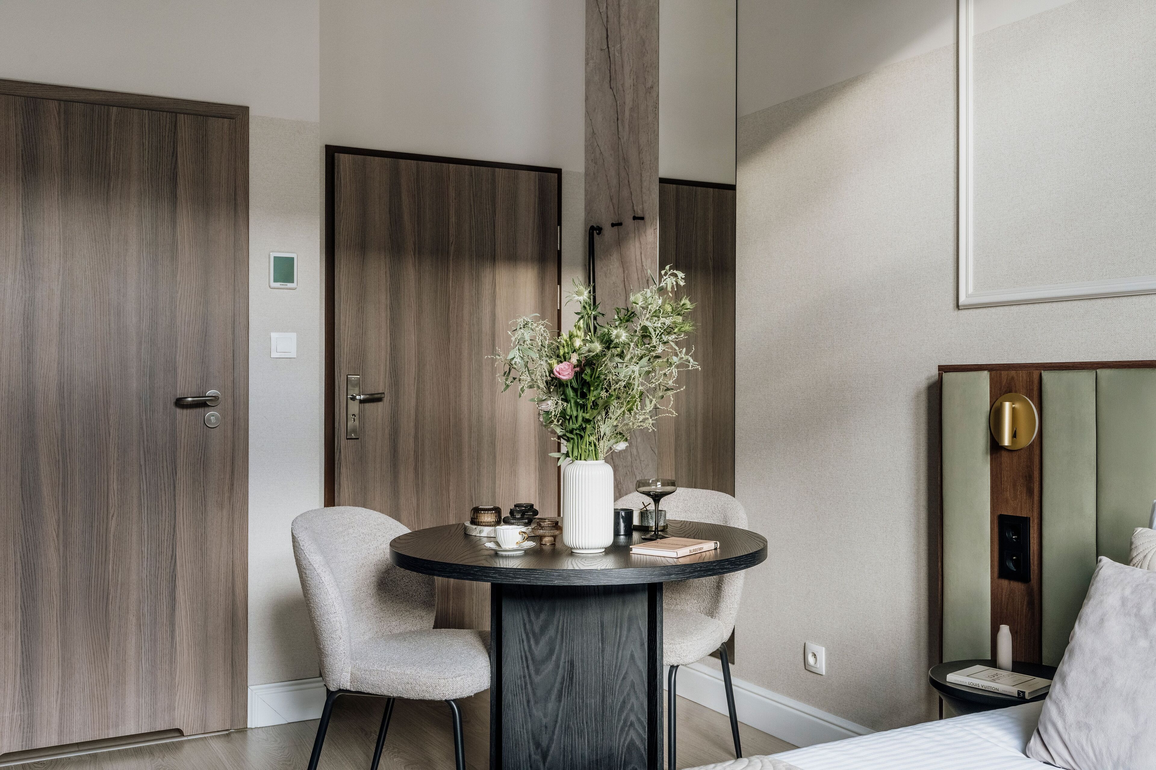 A modern hotel room with a round dining table, two chairs, a vase with flowers, a small side table with a book and a lamp, and a bed with a headboard.