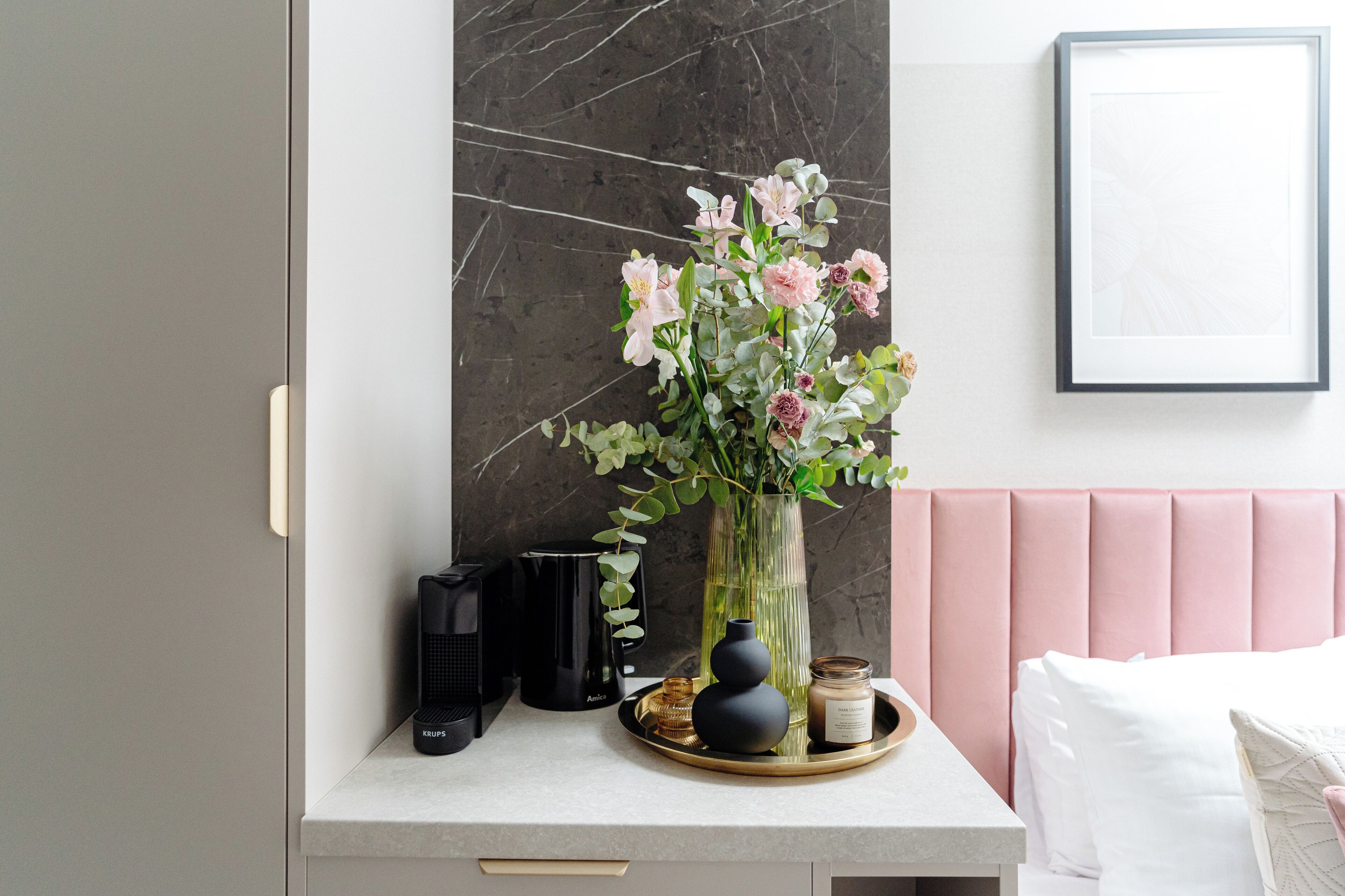 Un dormitorio moderno con pared de mármol, cabecero rosa y mesita de noche con un jarrón de flores, una cafetera y una bandeja con una vela y un tarro.