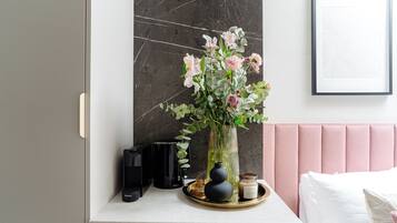 Un dormitorio moderno con pared de mármol, cabecero rosa y mesita de noche con un jarrón de flores, una cafetera y una bandeja con una vela y un tarro.