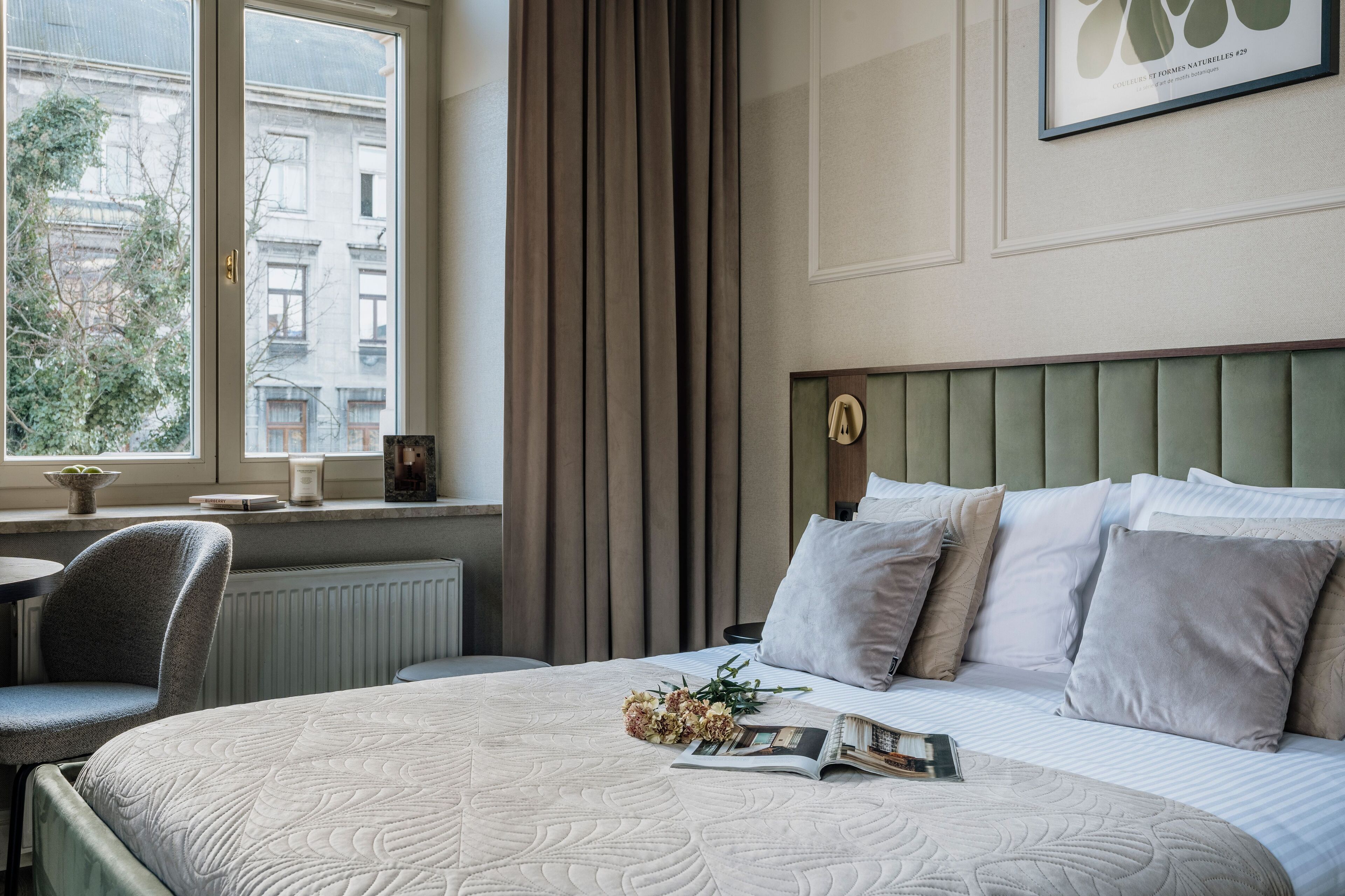 A neatly arranged hotel room with a large bed, a bedside table with a lamp, a chair, and a window offering a view of buildings outside.