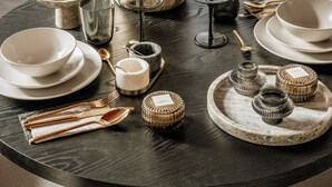 Een ronde eettafel gedekt met borden, kommen, glazen, bestek en een bloevas.