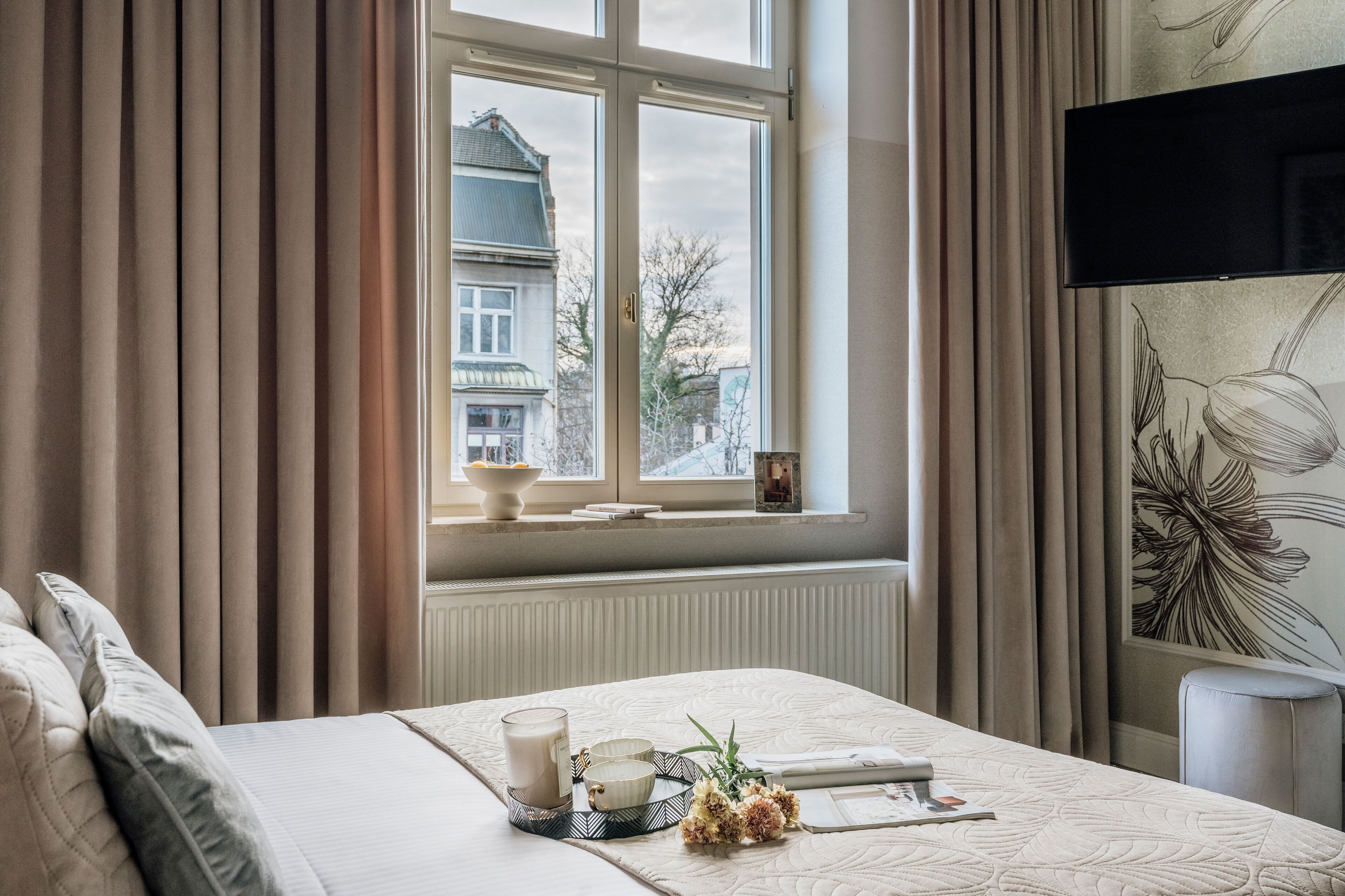 Hotellihuone, jossa on sänky, ikkuna verhoilla, patteri ja seinälle asennettu taulutelevisio.
